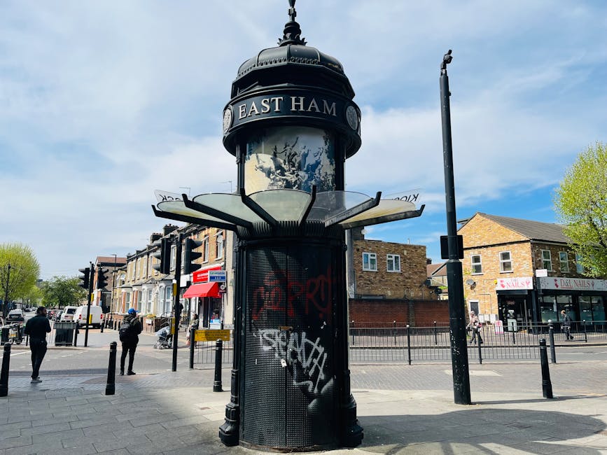 A historic cylindrical street clock with a domed top displaying the words 'EAST HAM' in white letters, situated at a busy intersection in Southfields SW18. The clock features a glass enclosure with de