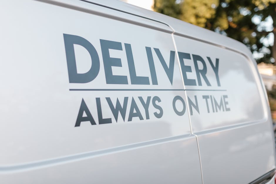 A close-up of the side of a white delivery van with the words 'DELIVERY ALWAYS ON TIME' printed in bold, dark grey letters. The van is parked outdoors in a well-lit environment, with a background of blurred trees and sky. Inside the cargo area, various packing materials such as cardboard boxes, plastic wrapping, and fabric covers are visible, along with furniture pieces including a wooden chair and a small table. Several items are being carefully loaded into the van using equipment like trolleys and straps, indicating a professional home relocation process carried out by Man With a Van Southfields. The image captures the loading activity in a residential setting, supporting services such as removals and furniture transport, reflecting efficient packing and moving procedures suited for house removals in Southfields SW18.