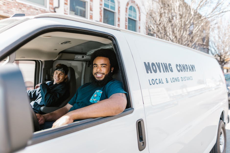 Inside the cab of a white moving van registered with Man With a Van Southfields, showing two smiling men seated in the front seats during a home relocation. The man in the driver’s seat has a dark curly hairstyle and a beard, wearing a dark blue t-shirt, while the passenger, a woman with short dark hair and a headband, is dressed in a black jacket with white accents. The van's interior reveals a clean, utilitarian space with a dashboard and steering wheel visible through the open window. The exterior of the van features professional branding for a local and long-distance removals service, indicating a furniture transport or packing and moving process. In the background, residential buildings and leafless trees suggest an urban setting, with natural daylight illuminating the scene, supporting the logistics of house removals for clients in Southfields SW18. Man With a Van Southfields specializes in efficient loading, packing, and transportation of household items, ensuring a smooth house move.