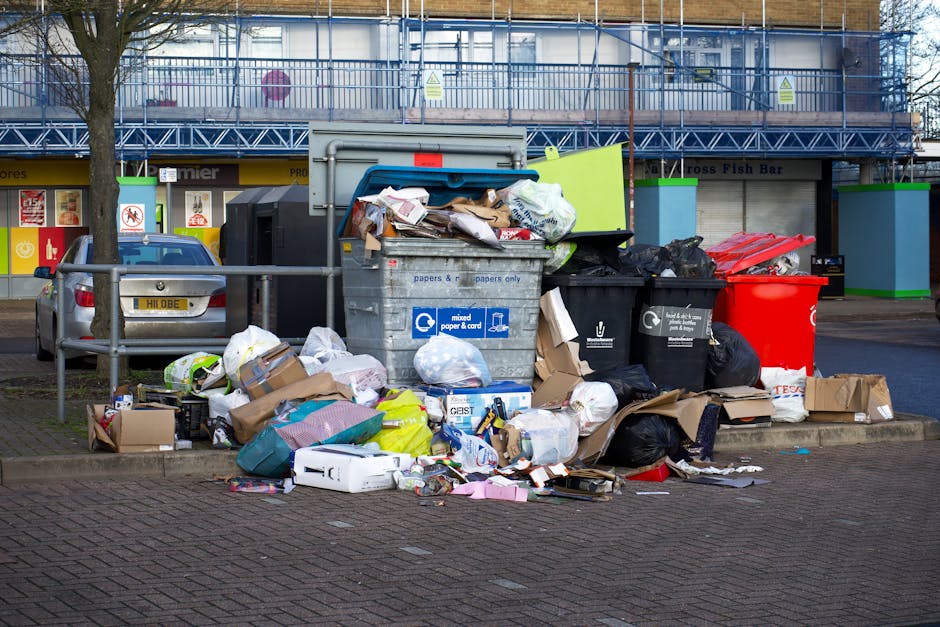 Outdoor scene showing overflowing waste and recycling bins placed on a paved sidewalk, with various black, red, and green rubbish bags, cardboard boxes, and paper waste piled around and on top of the bins. A parked silver car is situated nearby behind a metal railing, and behind the waste collection area, there are commercial or retail buildings with storefronts and signs. The setting appears to be in an urban environment during daylight hours, with a tree visible on the left side. The image captures the typical mess associated with waste disposal in a residential or commercial area, relevant to moving and packing logistics when disposing of unwanted items as part of home relocation services offered by companies like Man With a Van Southfields.