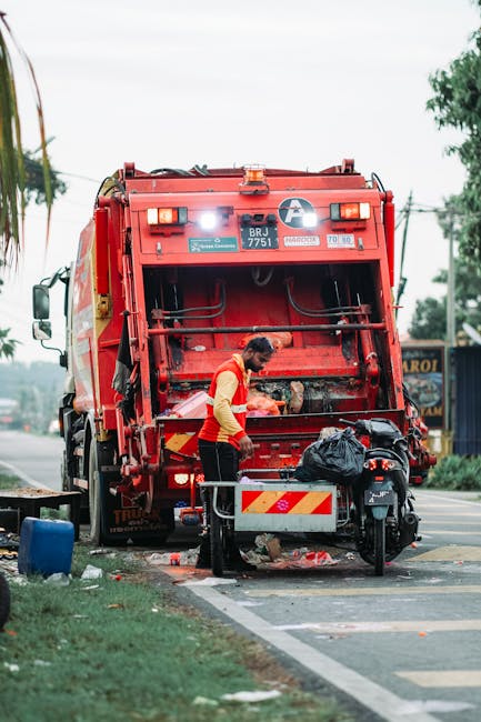 A worker wearing a yellow and red high-visibility jacket is loading or unloading rubbish from a red waste collection truck parked on the side of a street. The truck's rear hatch is open, revealing the interior where the worker appears to be handling black plastic bags and other waste materials. The scene includes a small baggage cart or trolley positioned nearby, and the street environment features a sidewalk, a blue container, and a commercial building in the background. Overhead, utility lines and trees are visible, with natural daylight illuminating the scene. This image reflects the process of waste collection and disposal, which relates to council bulky waste rules in SW18, and may be part of the logistical considerations for home relocation or moving services offered by Man With a Van Southfields, especially when managing unwanted items during a move.