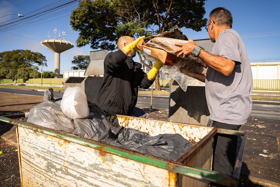 A worker wearing a yellow and red high-visibility jacket is loading or unloading rubbish from a red waste collection truck parked on the side of a street. The truck's rear hatch is open, revealing the interior where the worker appears to be handling black plastic bags and other waste materials. The scene includes a small baggage cart or trolley positioned nearby, and the street environment features a sidewalk, a blue container, and a commercial building in the background. Overhead, utility lines and trees are visible, with natural daylight illuminating the scene. This image reflects the process of waste collection and disposal, which relates to council bulky waste rules in SW18, and may be part of the logistical considerations for home relocation or moving services offered by Man With a Van Southfields, especially when managing unwanted items during a move.