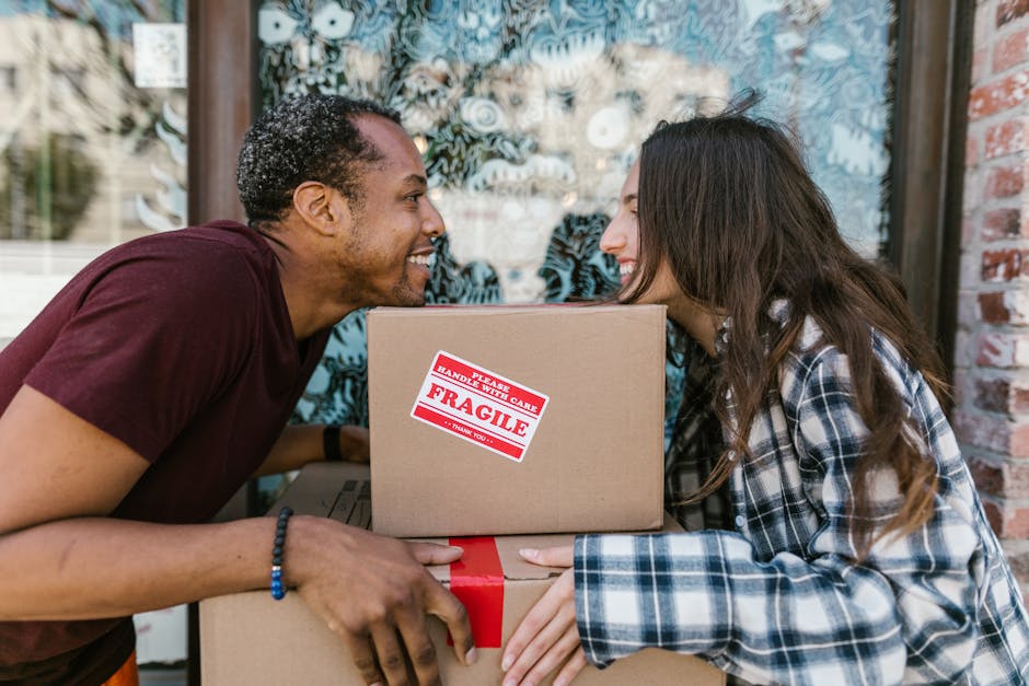 A man with short, curly dark hair and a woman with long, wavy brown hair smiling faces closely as they exchange a cardboard box during a home relocation process outside a brick building with large windows decorated with patterned curtains. The man, dressed in a maroon t-shirt and wearing a beaded bracelet on his wrist, supports a box labeled 'Fragile' with red and white handling care stickers, indicating careful packing for moving and furniture transport. The woman, wearing a plaid shirt, holds another box with her hand, assisting in loading or unloading. They are positioned near a doorway or driveway, with apparent natural lighting highlighting the task of packing and moving. The scene captures the collaborative effort involved in house removals, with the focus on the careful handling of packed items for a professional relocation service by Man With a Van Southfields.