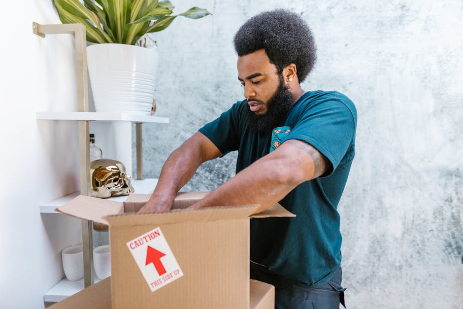 A man with dark, curly hair and a beard is standing inside a home, looking into an open cardboard box with a red and white caution sticker that reads 'CAUTION THIS SIDE UP.' He is wearing a dark teal T-shirt and is carefully placing items into the box, engaged in packing for a home relocation. In the background, there is a white wall with a shelving unit holding decorative objects, including a gold skull sculpture, ceramic cups, and a potted plant with long green leaves. The scene is illuminated by natural light, emphasizing the organized interior environment. This image illustrates the packing and loading process involved in house removals, highlighting careful handling of belongings in preparation for furniture transport by a professional removals service like Man With a Van Southfields.