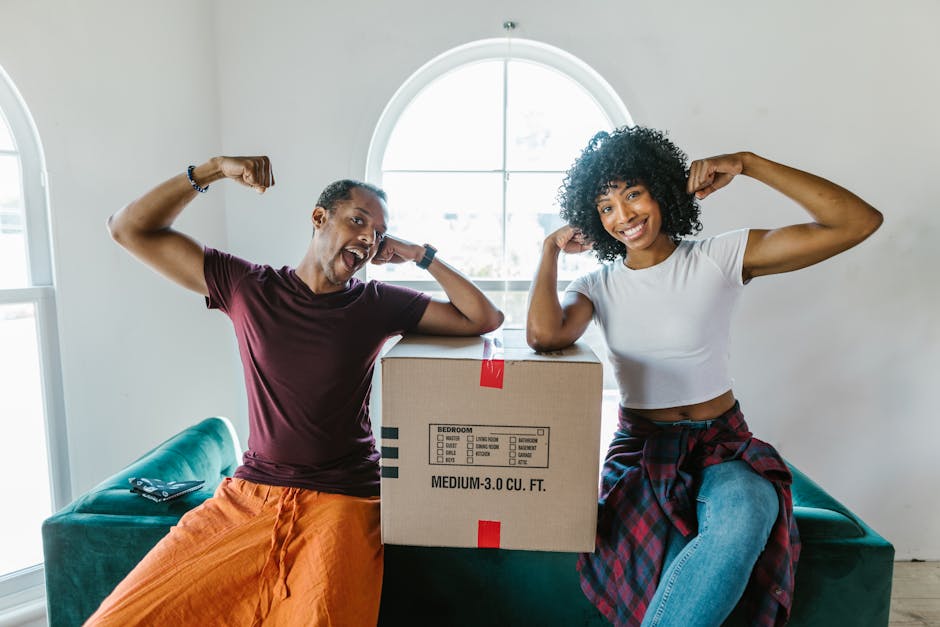 A man with short, curly dark hair and a woman with long, wavy brown hair smiling faces closely as they exchange a cardboard box during a home relocation process outside a brick building with large windows decorated with patterned curtains. The man, dressed in a maroon t-shirt and wearing a beaded bracelet on his wrist, supports a box labeled 'Fragile' with red and white handling care stickers, indicating careful packing for moving and furniture transport. The woman, wearing a plaid shirt, holds another box with her hand, assisting in loading or unloading. They are positioned near a doorway or driveway, with apparent natural lighting highlighting the task of packing and moving. The scene captures the collaborative effort involved in house removals, with the focus on the careful handling of packed items for a professional relocation service by Man With a Van Southfields.