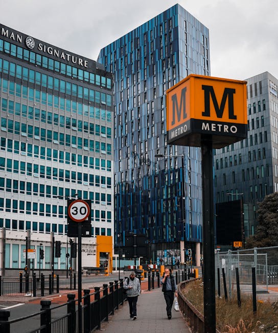 A busy urban scene showing modern office buildings with glass and steel exteriors, including the Man Signature building and other high-rise structures, situated near Southfields station in London. In