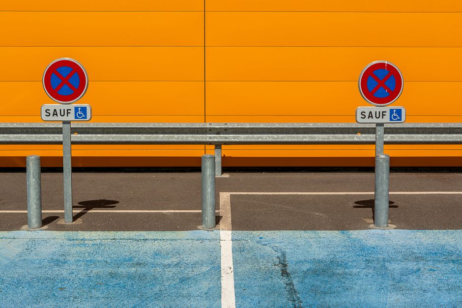 Two metal posts with no parking signs mounted on top, located on a paved surface in front of a orange wall. The signs feature a red circle with a blue background and a red cross, indicating no parking, with additional small blue disability access signs below labeled 'SAUF'. The area includes a metal barrier extending between the posts and a white no-parking line painted on the asphalt. Behind the posts, the pavement is marked with a designated loading bay area in blue, with visible markings for vehicle loading and unloading. The scene is well-lit with natural daylight and appears to be part of a designated space for moving or home relocation activities, aligning with house removals and furniture transport services provided by Man With a Van Southfields, situated adjacent to a building involved in relocation logistics.