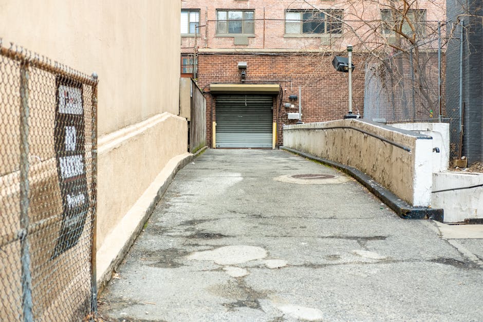 A paved alleyway leading to a garage with a closed metallic roller shutter door, situated behind a residential building in Southfields. On the left side of the alley, a beige concrete wall runs along the property, and a chain-link fence with a small electrical meter box attached is visible. To the right, a white concrete barrier with metal railings separates the driveway from an adjacent area, showing signs of weathering and slight staining. The surface of the alley shows patches of different shades of asphalt and some visible cracks and wear, indicating use and age. In the background, there is a brick building with multiple windows, and a security camera and outdoor lighting fixtures are mounted on the wall. The scene suggests a location used for furniture transport or home relocation activities, potentially managed by Man With a Van Southfields, with the alley serving as part of a loading area for moving services and parking regulations relevant to Southfields.
