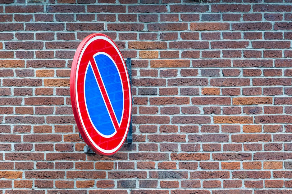 Two metal posts with no parking signs mounted on top, located on a paved surface in front of a orange wall. The signs feature a red circle with a blue background and a red cross, indicating no parking, with additional small blue disability access signs below labeled 'SAUF'. The area includes a metal barrier extending between the posts and a white no-parking line painted on the asphalt. Behind the posts, the pavement is marked with a designated loading bay area in blue, with visible markings for vehicle loading and unloading. The scene is well-lit with natural daylight and appears to be part of a designated space for moving or home relocation activities, aligning with house removals and furniture transport services provided by Man With a Van Southfields, situated adjacent to a building involved in relocation logistics.