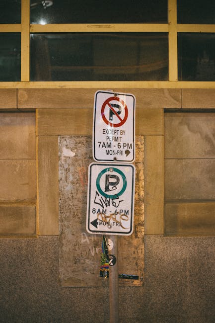 Photograph of a vertical metal parking restriction sign mounted on a wall adjacent to a window with yellow framing, displaying two sections of parking and loading regulations. The top section indicates no parking except by permit from 7am to 6pm Monday to Friday, and the bottom section permits parking with a valid permit from 8am to 6pm on weekdays. The signs are attached to a metal post, which has some small attached objects hanging on it, with a background of a stone wall and a window with yellow framing. This image may be relevant in the context of house removals or relocation services provided by Man With a Van Southfields, particularly when considering parking permits and restrictions during home or furniture transport and loading processes.