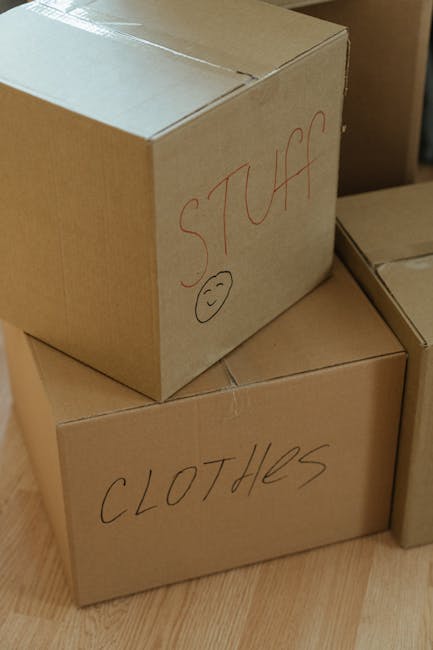 Two cardboard boxes are stacked indoors on a wooden floor, with the larger box on the bottom and a smaller one on top. The larger box is labeled 'CLOTHES' in black marker, while the smaller box is lab