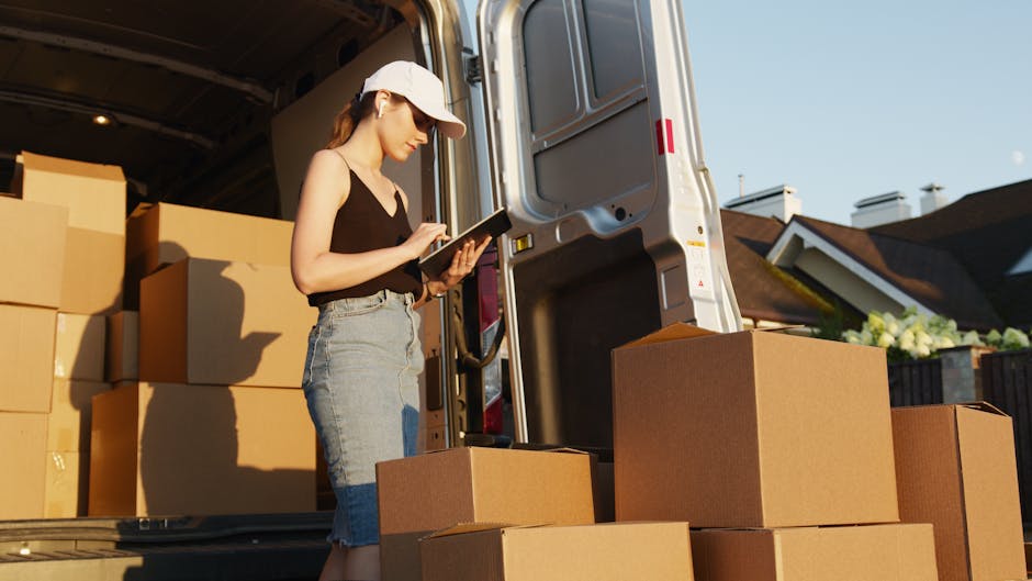 A woman wearing a beige cap, black sleeveless top, and light blue jeans stands beside an open moving van during a home relocation task. She is holding a tablet device in her hands and appears to be checking inventory or coordinating the packing process. Inside the van, several large cardboard boxes are stacked, along with packing materials such as plastic wrap and protective blankets. Some boxes are placed on the pavement outside the vehicle, ready for loading or unloading. The van is parked in an outdoor residential area, with nearby houses and a clear sky in the background. The scene reflects the process of furniture transport and packing during a professional removals service, emphasizing careful handling and organized logistics. Man With a Van Southfields, a company specializing in removals, is involved in this home moving activity, ensuring efficient and secure furniture transport.