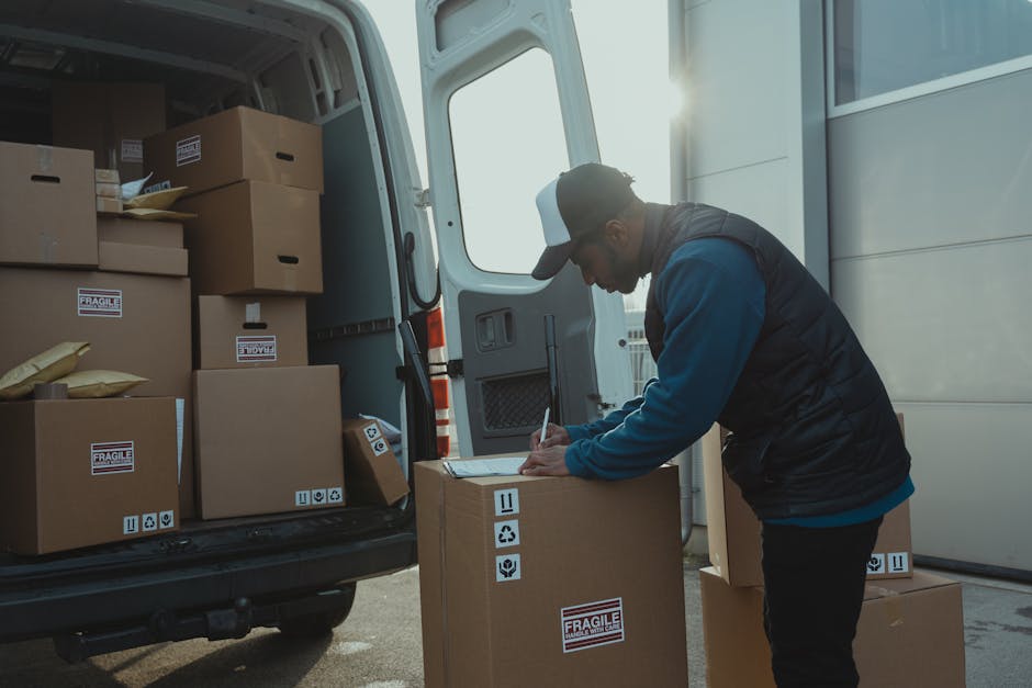 A woman wearing a beige cap, black sleeveless top, and light blue jeans stands beside an open moving van during a home relocation task. She is holding a tablet device in her hands and appears to be checking inventory or coordinating the packing process. Inside the van, several large cardboard boxes are stacked, along with packing materials such as plastic wrap and protective blankets. Some boxes are placed on the pavement outside the vehicle, ready for loading or unloading. The van is parked in an outdoor residential area, with nearby houses and a clear sky in the background. The scene reflects the process of furniture transport and packing during a professional removals service, emphasizing careful handling and organized logistics. Man With a Van Southfields, a company specializing in removals, is involved in this home moving activity, ensuring efficient and secure furniture transport.